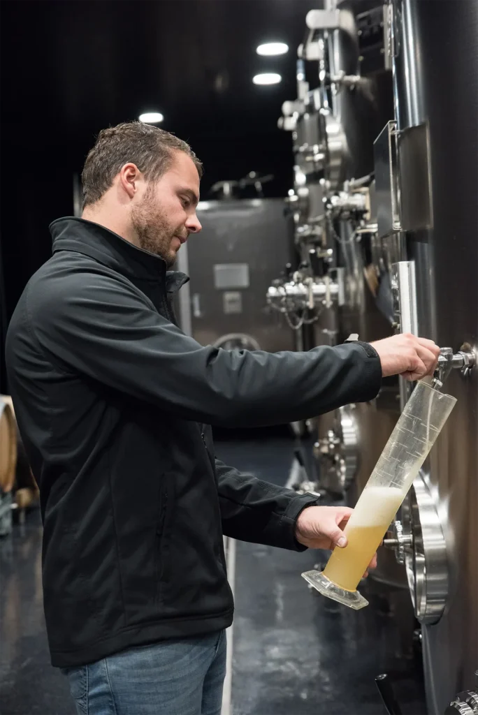 Dans la cave en inox, un homme prélève une cuvée au robinet d’une cuve pour contrôler la dégustation et le suivi du champagne artisanal.